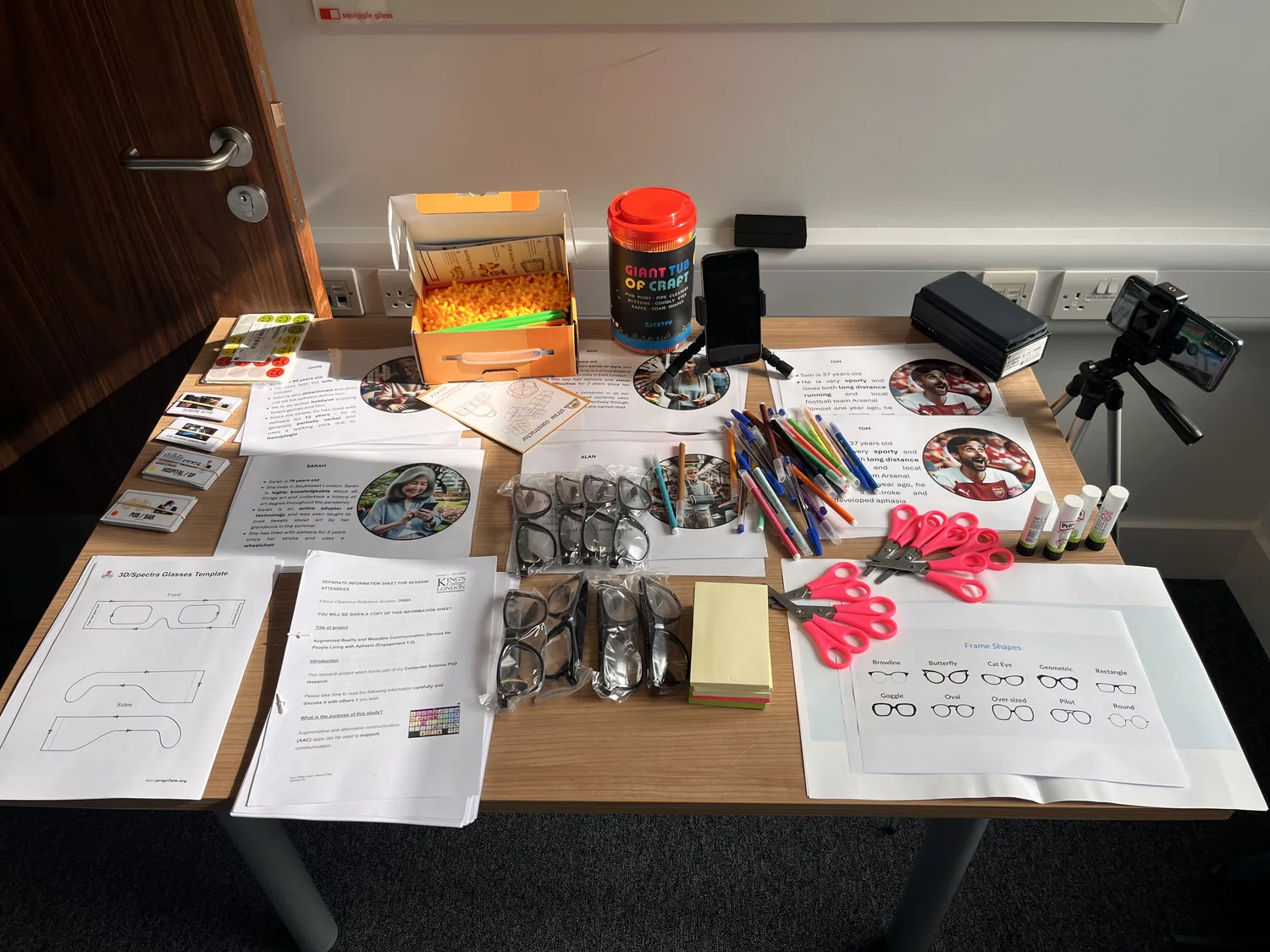 A workshop table prepared with cardboard glasses, character prompts, pens, frame templates and construction materials