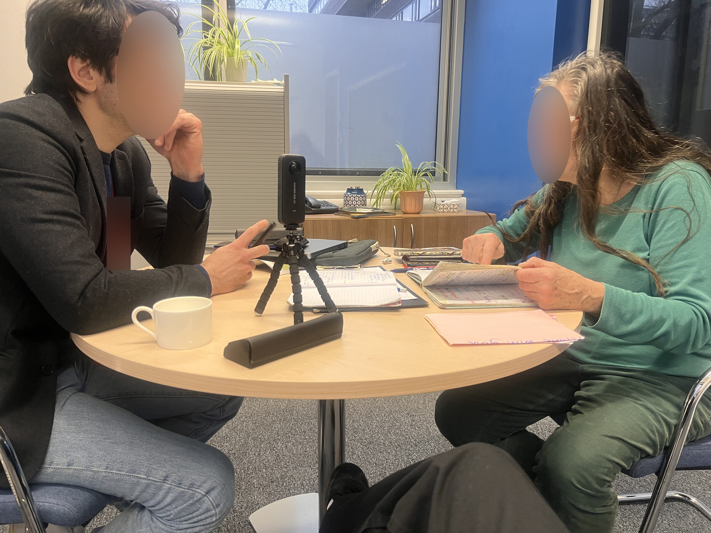 Researcher and participant seated at a round table, diary open between them and a video camera on a tripod recording the session